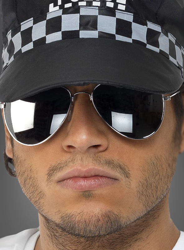 Eine Person, die eine verspiegelte „Polizei“-Sonnenbrille und eine schwarze Mütze mit Karomuster trägt, blickt direkt in die Kamera und zeigt dabei einen neutralen Gesichtsausdruck.