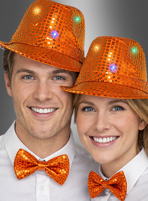 Zwei Personen, die leuchtend orangefarbene Trilby-Hüte mit LED und dazu passende orangefarbene Pailletten-Fliegen tragen und vor einem grauen Hintergrund in die Kamera lächeln.