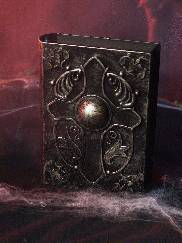 Ein dunkles, kunstvoll verziertes „Zauberbuch“-Deko-Animatronic mit Licht und Sound, das sich durch geprägte Motive und eine zentrale Metallkuppel auszeichnet, ruht inmitten von Spinnweben und rotem Licht – und sorgt so für eine bezaubernde Halloween-Dekoration.