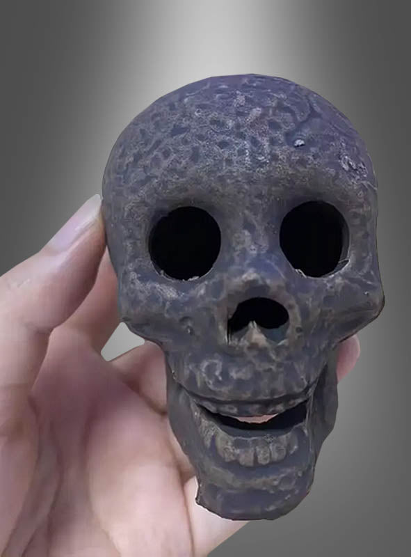 Eine Hand hält einen kleinen, dunklen, verwitterten, feuerfesten Keramik-Totenkopf mit großen Augenhöhlen und einem leicht geöffneten Mund vor einem grau verlaufenden Hintergrund – ein faszinierendes Halloween-Dekostück.