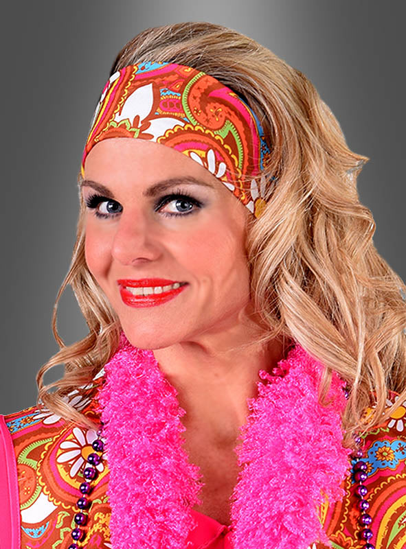 Eine Frau mit welligem blondem Haar trägt das farbenfrohe Hippie-Stirnband „Martina“, ein gemustertes Oberteil mit Paisley-Print und eine leuchtend pinkfarbene Federboa und lächelt vor einem grauen Hintergrund in die Kamera.