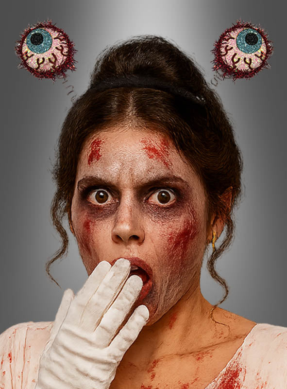 Eine Frau mit Zombie-Make-up und weißen Handschuhen hält sich vor Schreck die Hand vor den Mund. Sie trägt den „Lustiger Augen Haarreif 2D Halloween“, der ihrem gruseligen Look eine verspielte Note verleiht, da zwei illustrierte, blutunterlaufene Augen über ihrem Kopf schweben.