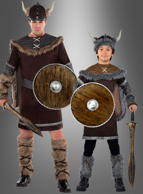 Ein Erwachsener und ein Kind, als Wikinger verkleidet, stehen vor einem grauen Hintergrund mit Farbverlauf. Beide halten jeweils ein „Rundschild 40 cm in Holzoptik“ – authentische runde Schilde im Holzlook, die von traditionellen Rundschild-Designs inspiriert sind – sowie Schwerter in der Hand; beide tragen Hörnerhelme und mit Kunstfell besetzte Kostüme.