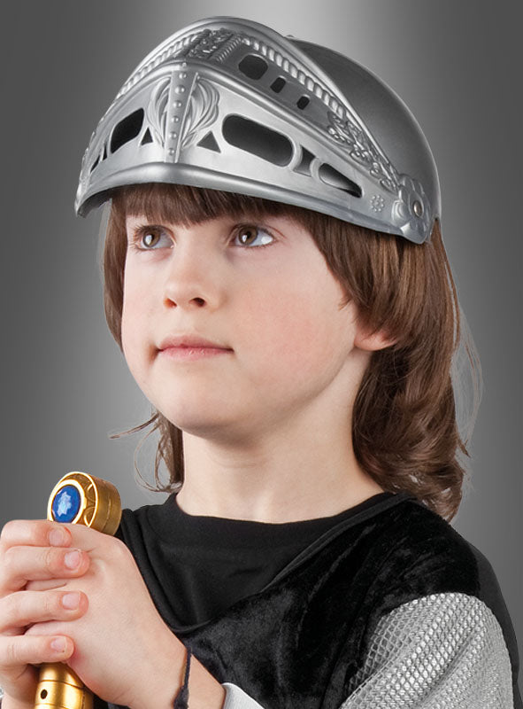 Ein kleines Kind, das einen silbernen Ritterhelm für Kinder trägt und ein goldenes Zepter in der Hand hält, blickt mit ausdruckslosem Gesicht vor einem grauen Hintergrund mit Farbverlauf nach oben.