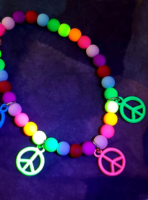 Das „Bunte Hippiearmband mit Peace-Zeichen“ ist ein einzigartiges Hippie-Schmuckstück, das mit bunten Perlen und drei Anhängern in Form von Peace-Zeichen verziert ist und eindrucksvoll vor einem dunklen Hintergrund fotografiert wurde.
