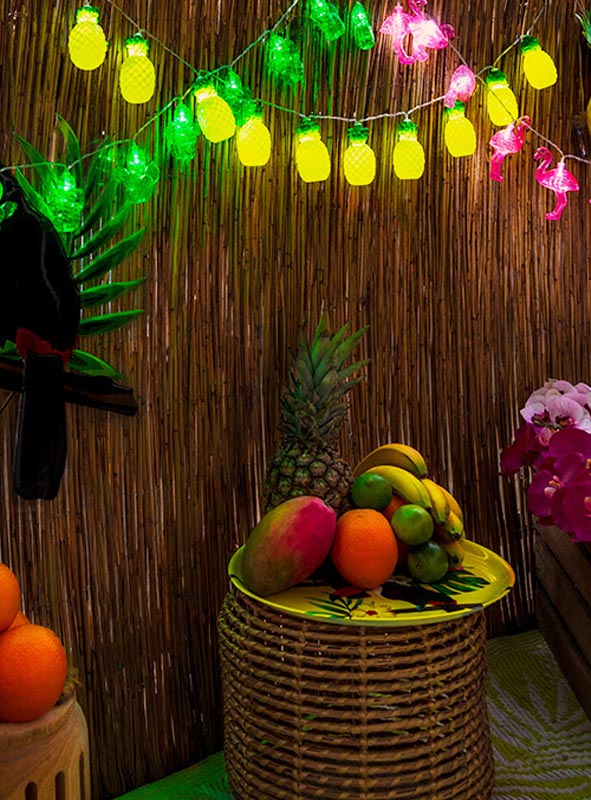 Auf einem Korb-Tisch sind verschiedene tropische Früchte unter der Lichterkette „Ananas“ 140 cm „Sommerparty“ ausgestellt, während rosa Blumendekorationen die festliche „Sommerparty“-Dekoration vor einer Bambuswand unterstreichen – perfekt für jede Hawaii-Party.