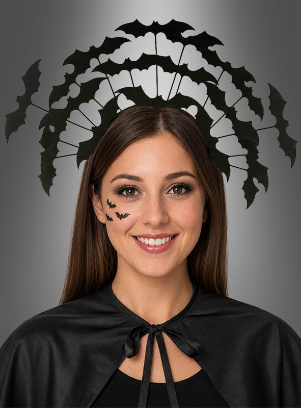 Eine Frau lächelt vor einem grauen Hintergrund. Sie trägt einen schwarzen Umhang, Fledermaus-Gesichtssticker und den Haarreif „Fledermaus-Schwarm“ – ein perfektes Halloween-Kostüm-Accessoire für jede gruselige Feier.