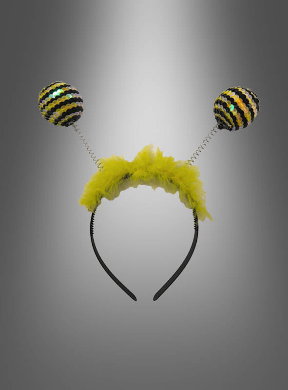 Ein „Bienen-Fühler“-Haarreif mit Glitzer, mit gelbem, flauschigem Rand und zwei federnd befestigten, schwarz-gelb gestreiften Pompons, die Bienenantennen ähneln – perfekt als Zubehör für ein Bienenkostüm –, präsentiert vor einem grauen Hintergrund mit Farbverlauf.