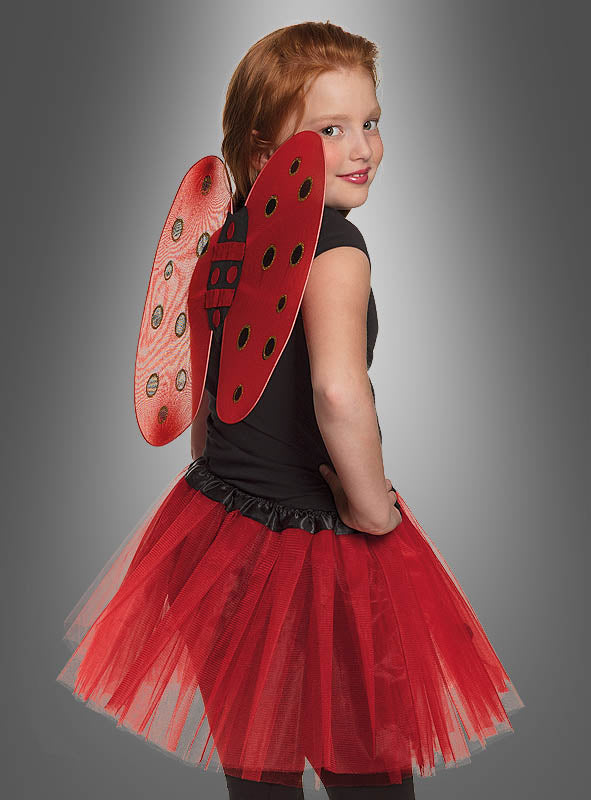 Ein Kind, das ein schwarzes Shirt, ein rotes Tutu und das „Marienkäfer“-Flügel-Set für Kinder trägt, steht mit den Händen in die Hüften gestützt vor einem grauen Hintergrund und blickt über die Schulter – ein bezauberndes Karnevalskostüm für Kinder, das sich perfekt für jeden festlichen Anlass eignet.