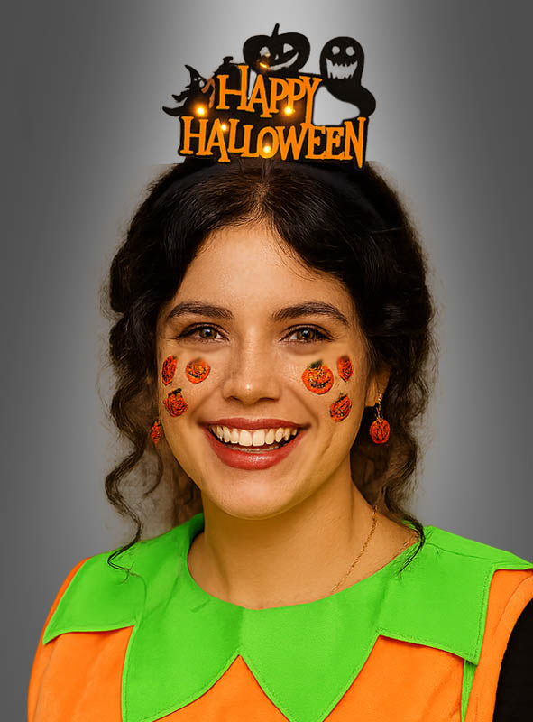 Eine Frau lächelt, während sie das Stirnband „Happy Halloween Haarreif mit Licht“ mit Kürbis- und Geistermotiven trägt, dazu Kürbis-Ohrringe, Aufkleber mit Kürbisköpfen und ein grün-orangefarbenes Kostüm – perfekt als lustiges Halloween-Accessoire.