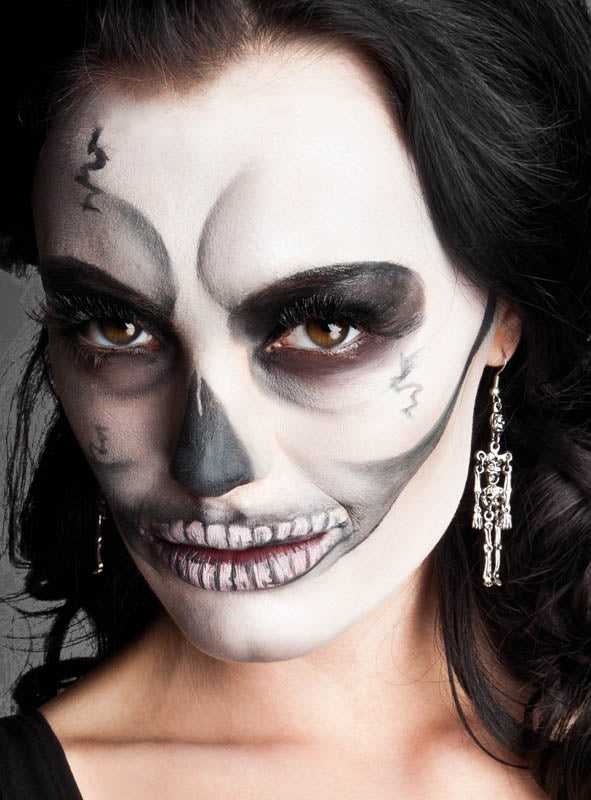 Eine Person mit langen dunklen Haaren, die eine Skelett-Gesichtsbemalung und auffällige Ohrringe sowie Skelett-Halloween-Schmuck trägt und direkt in die Kamera blickt – eine markante Wahl für einen gruseligen Look.