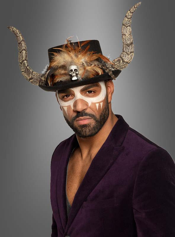 Ein Mann in einem violetten Blazer trägt den „Voodoo-Zylinder mit Hörnern Papa Legba“, der mit Federn und einem Totenkopf verziert ist; sein Gesicht ist weiß und braun bemalt und weist auf grauem Hintergrund Reißzähne auf.