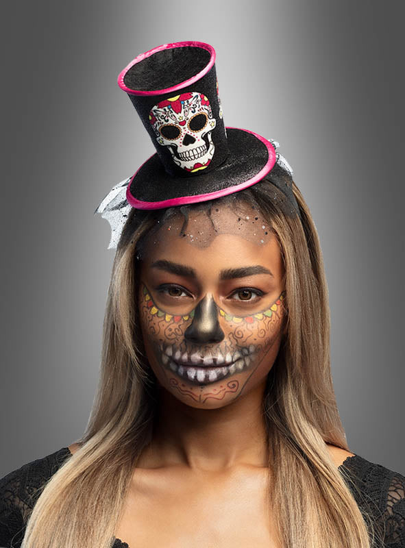 Eine Person, die einen kleinen „Tag der Toten“-Mini-Zylinder trägt und deren Gesicht mit aufwendigen Totenkopf- und Blumenmotiven im Stil des „Tag der Toten“ bemalt ist, steht vor einem neutralen Hintergrund.