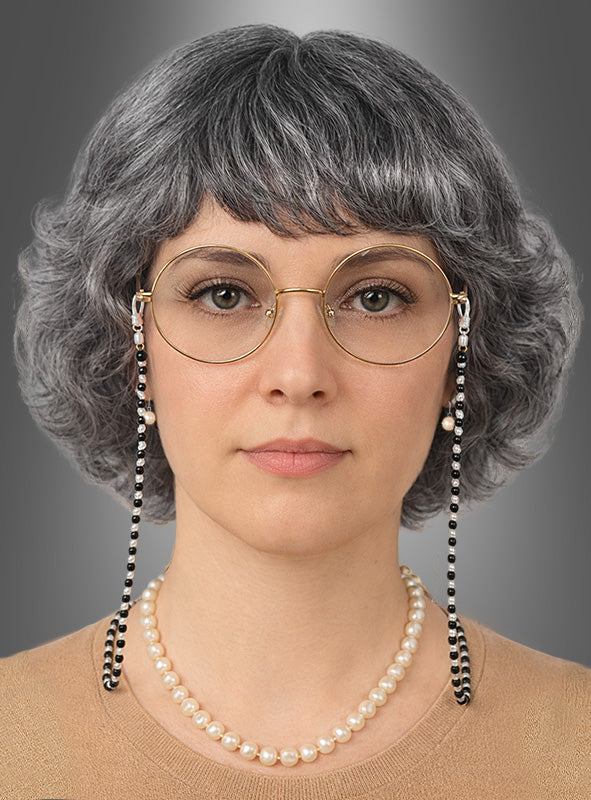 Eine Frau mit kurzem, grauem Haar, das als Perücke aus dem 4-teiligen „Oma-Perücken-Set“ gestylt ist, trägt eine runde Brille an einer Kette, ein beiges Oberteil und eine Perlenkette aus einem „Oma-Schmuck-Set“ und blickt vor einem grauen Hintergrund mit Farbverlauf in die Kamera.