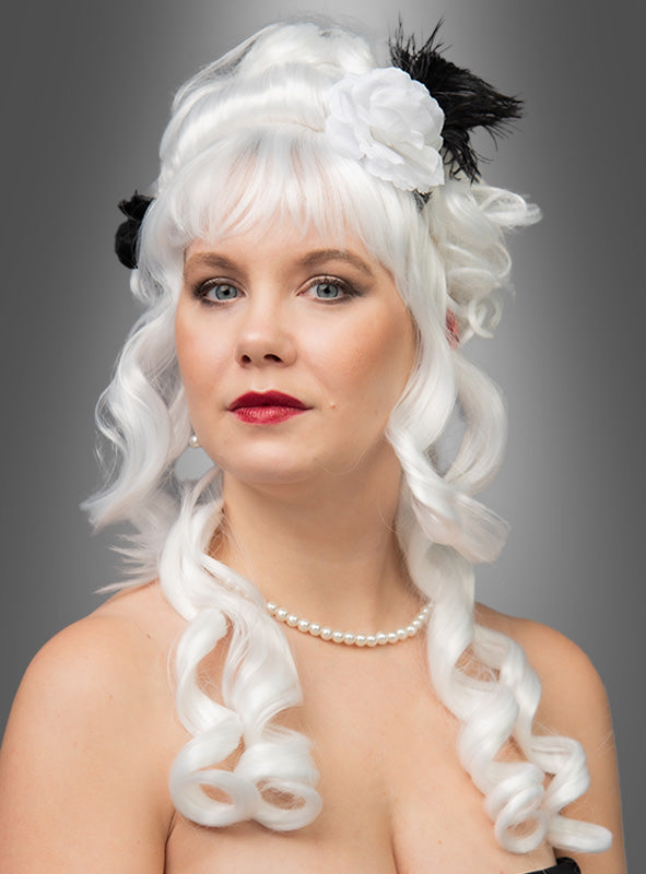 Eine Frau mit blasser Haut, die die „Deluxe Barock Perücke Marie Antoinette“ trägt – eine weiße, lockige Perücke, verziert mit einer weißen Blume und einer schwarzen Feder –, dazu eine Perlenkette, vor einem grauen Hintergrund.