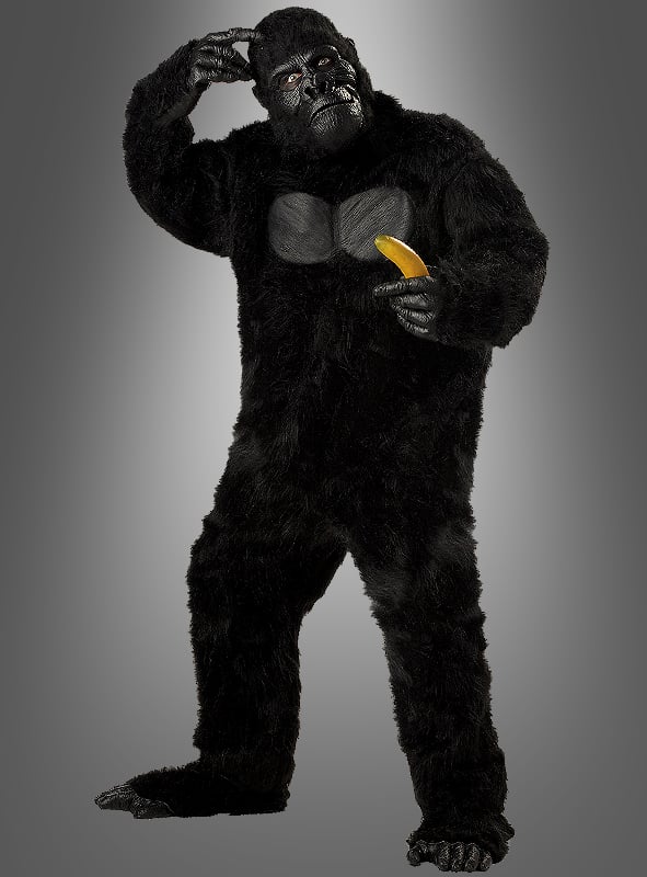 Vor einem grauen Hintergrund steht eine Person, die ein XXL-Gorilla-Kostüm in Übergröße trägt, auf einem Bein, kratzt sich mit einer Hand am Kopf und hält in der anderen Hand eine Banane.
