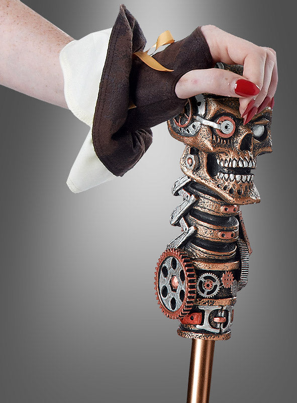 Eine Person in einem braun-weißen Steampunk-Kostüm hält vor einem grauen Hintergrund den „Gehstock Steampunk Alien“ in der Hand – einen Spazierstock, dessen Kopfstück aus einem metallischen, im Steampunk-Stil gestalteten Totenkopf mit Zahnrädern besteht –, was diesen einzigartigen Gehstock zum perfekten Spazierstock-Accessoire macht.