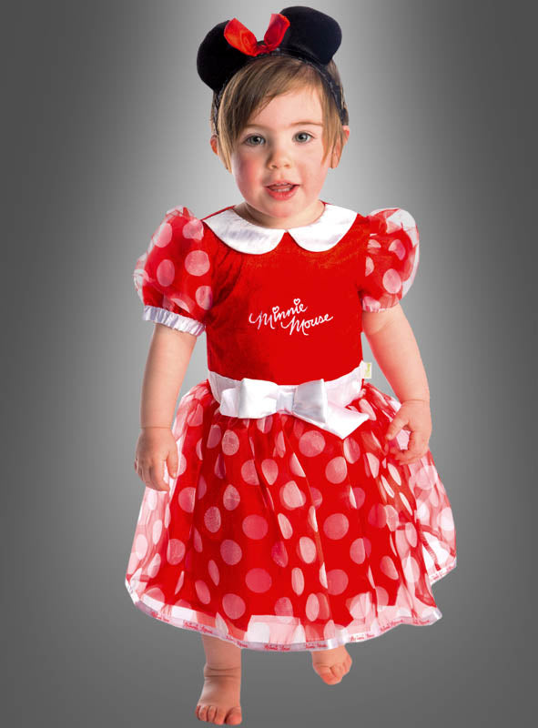 Ein kleines Kind steht barfuß da und trägt ein bezauberndes Minnie-Maus-Kleid aus rotem Stoff mit weißen Punkten sowie ein passendes schwarzes Stirnband mit Mausohren, das mit einer roten Schleife verziert ist. Im Hintergrund ist ein grauer Farbverlauf zu sehen.
