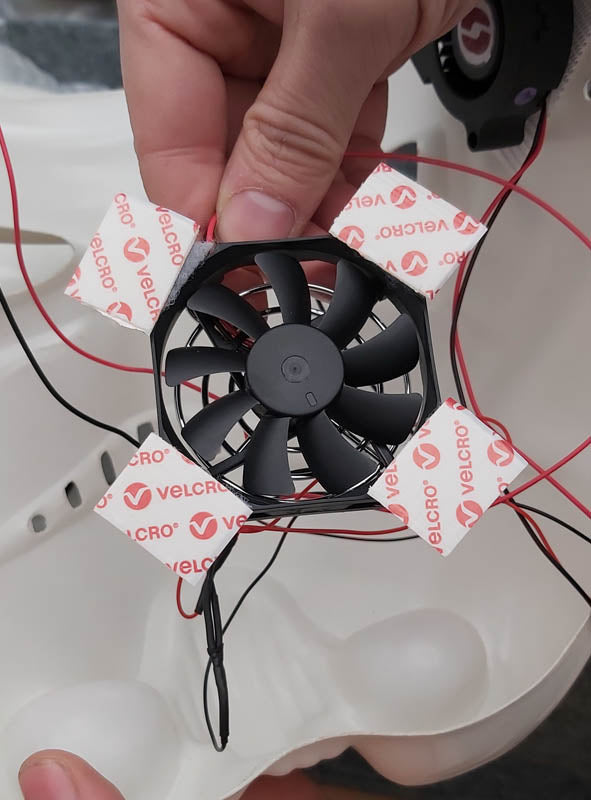Eine Hand hält einen kleinen schwarzen Ventilator, der Teil des „Lüfter-Systems für Masken und Helme“ ist. Er ist mit Klettverschlussstreifen und roten Drähten in einem weißen Kunststoffgehäuse befestigt – ideal, um den Tragekomfort Ihrer Star-Wars-Kostüme oder Cosplay-Helme zu verbessern.