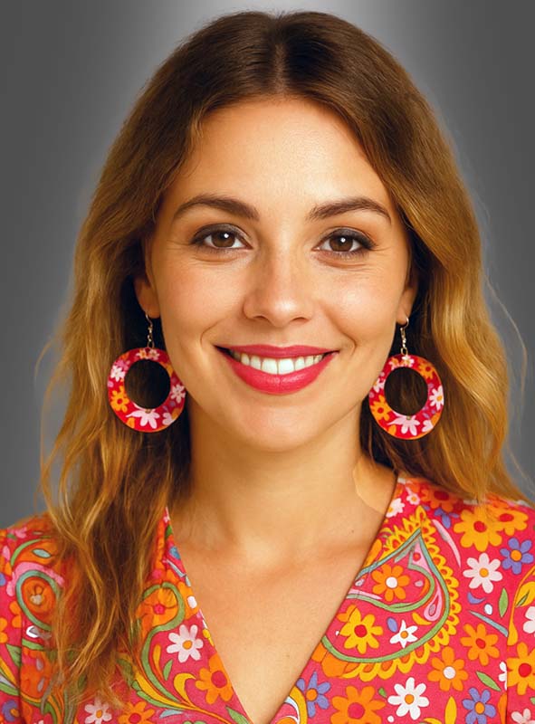 Eine Frau mit welligem braunem Haar trägt große runde Hippie-Ohrringe mit Blumenmotiven und ein farbenfrohes Oberteil mit Blumenmuster und lächelt vor einem grauen Hintergrund mit Farbverlauf in die Kamera.