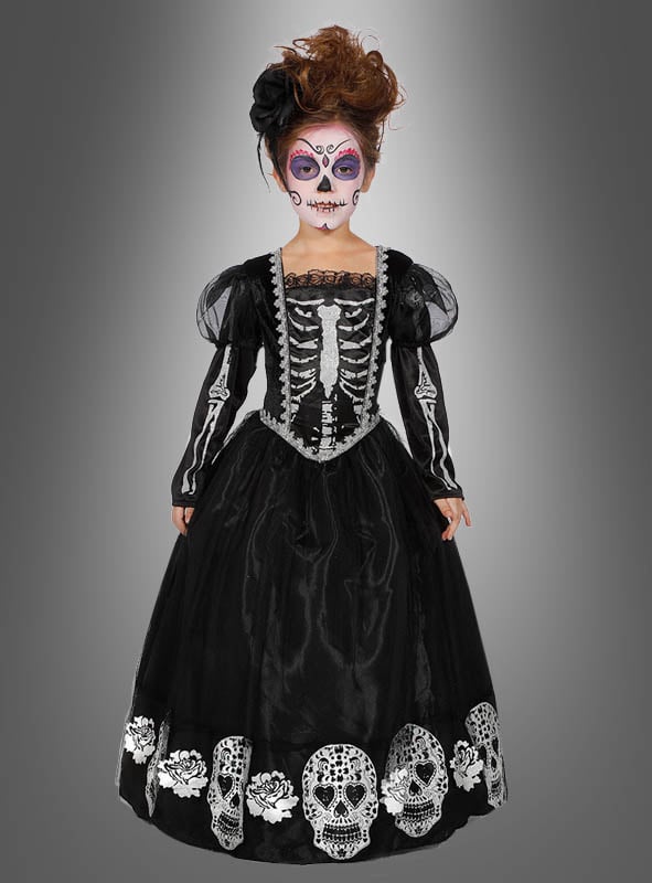 Ein Kind, das das „Deluxe Totenkopf-Kleid für Mädchen“ trägt – mit Skelettdetails und Totenköpfen am Saum – und eine von „Dia de los Muertos“ inspirierte Gesichtsbemalung trägt, steht vor einem grauen Hintergrund mit Farbverlauf.