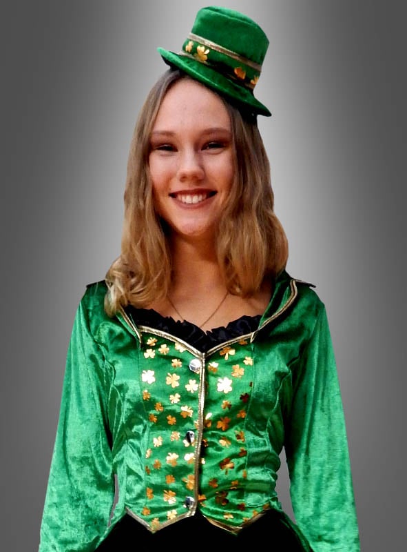 Vor einem grauen Hintergrund mit Farbverlauf steht eine lächelnde Person mit einem grünen Hut und einem grünen Samt-Outfit, das mit Kleeblättern verziert ist, und präsentiert das „Sexy Kobold“-Kostüm für Damen – perfekt für den St. Patrick’s Day.