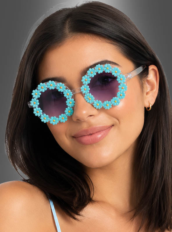 Eine Frau mit glattem, dunklem Haar präsentiert die „Hippie Blumen“-Sonnenbrille, die mit blauen und gelben Blumenverzierungen versehen ist, vor einem schlichten grauen Hintergrund.