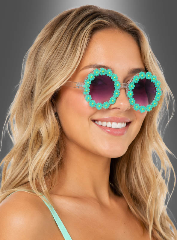 Eine Frau mit welligem blondem Haar lächelt vor einem grauen Hintergrund. Sie trägt die „Hippie Blumen“-Sonnenbrille – eine runde Sonnenbrille, die mit kleinen blauen Blumen verziert ist – und ein hellgrünes Oberteil.