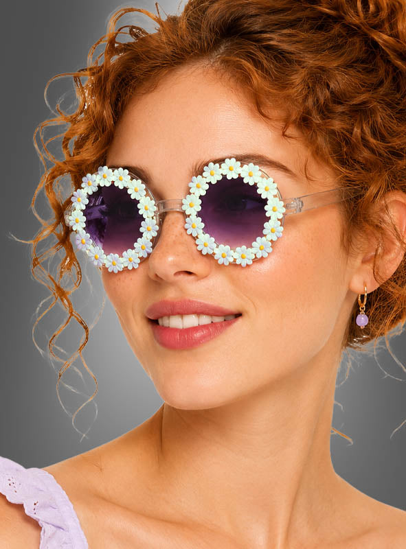Eine Frau mit lockigem rotem Haar trägt die „Hippie Blumen“-Sonnenbrille – eine runde Sonnenbrille, die mit künstlichen weißen Gänseblümchen verziert ist und violett getönte Gläser hat –, dazu kleine Creolen und ein lavendelfarbenes Oberteil, während sie leicht zur Seite blickt.