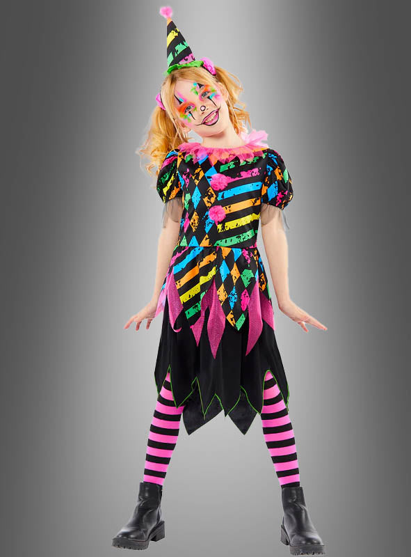 Ein Kind, das das „Zirkus Clown Neon Kostüm Mädchen“ trägt – mit gestreiften Strumpfhosen, einem farbenfrohen, gemusterten Kleid, einem Partyhut, Gesichtsbemalung und Stiefeln – steht vor einem grauen Hintergrund und eignet sich somit perfekt für Kinderfasching-Feiern.