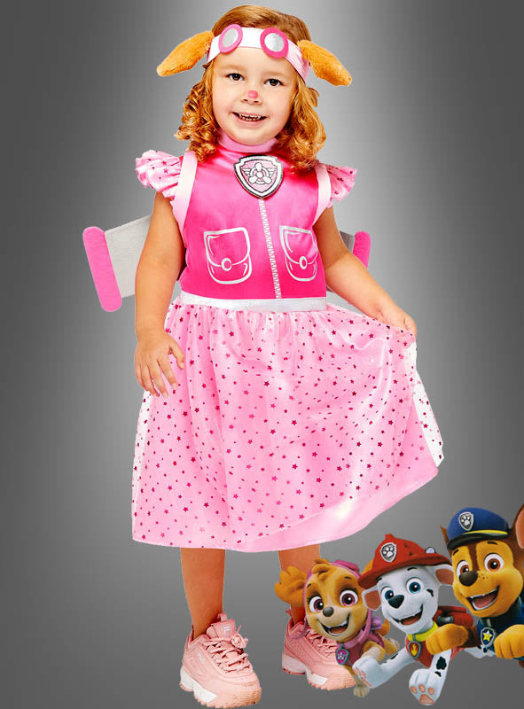 Ein junges Mädchen, das das „Skye Paw Patrol“-Kostüm für Mädchen (Deluxe-Ausführung) trägt, lächelt und hält ihr Kleid fest, auf dessen rechter unterer Ecke die Figuren aus der Zeichentrickserie „PAW Patrol“ abgebildet sind.
