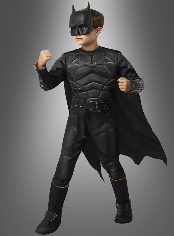 Ein Kind, das ein „Batman Luxus“-Kinderkostüm trägt – komplett mit Umhang, Maske und Handschuhen –, steht mit erhobenen Fäusten vor einem schlichten Hintergrund mit Farbverlauf.