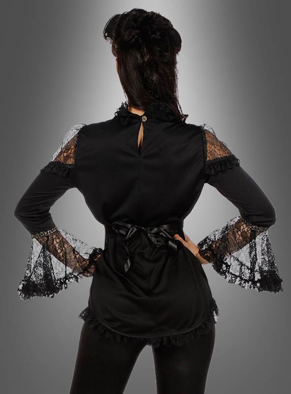 Eine Frau steht mit dem Rücken zur Kamera, die Hände in die Hüften gestemmt, und trägt die „Elegante Bluse mit Spitze, schwarz“ mit Rüschenverzierungen vor einem grauen Hintergrund mit Farbverlauf.
