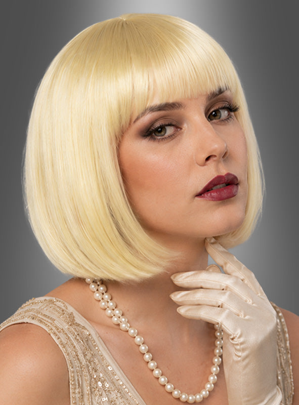 Eine Frau mit einer kurzen, blonden Bob-Frisur im Stil der 20er-Jahre (Charleston-Frisur), elegant gestylt, trägt eine Perlenkette, ein beiges Kleid und einen elfenbeinfarbenen Satinhandschuh und posiert mit der Hand am Kinn vor einem grauen Hintergrund.