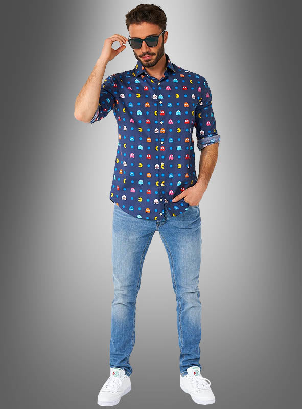 Ein Mann in einem „Pac-Man“-Hemd von OppoSuits im Retro-Stil einer 80er-Jahre-Mottoparty, hellblauen Jeans und weißen Sneakers, der eine Sonnenbrille trägt und vor einem grauen Hintergrund mit Farbverlauf posiert.