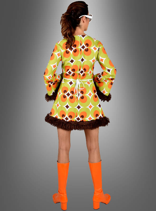 Eine Person in einem grün-braunen 70er-Jahre-Retro-Kleid für Damen mit grünen und braunen Retro-Mustern und braunen Fransen, orangefarbenen kniehohen Stiefeln und einer weißen Sonnenbrille – perfekt als 70er-Jahre-Damenkostüm – steht mit dem Rücken zur Kamera vor einem grauen Hintergrund mit Farbverlauf.