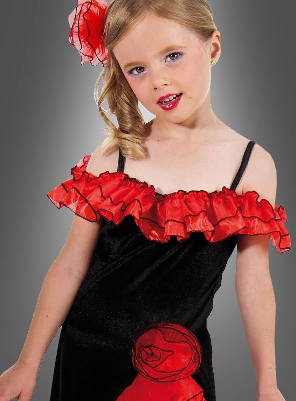 Ein junges Mädchen präsentiert das „Spanierin“-Kostüm für Kinder „Carmen“, bestehend aus einem schwarzen Kleid mit roten Rüschen und einem passenden roten Blumenaccessoire im Haar, vor einem grauen Hintergrund.
