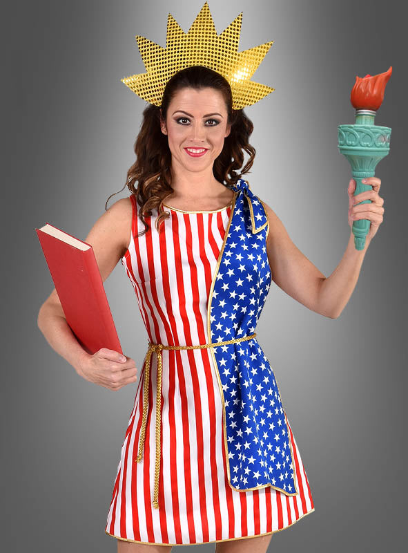 Eine Frau in einem von der Freiheitsstatue inspirierten „Stars & Stripes USA“-Flaggenkleid mit Sternen und Streifen, die ein rotes Buch und eine Fackel in der Hand hält und eine Krone mit Sonnenstrahlen trägt – dieses Kostüm eignet sich perfekt, um vor dem grauen Hintergrund ein patriotisches Statement zu setzen.