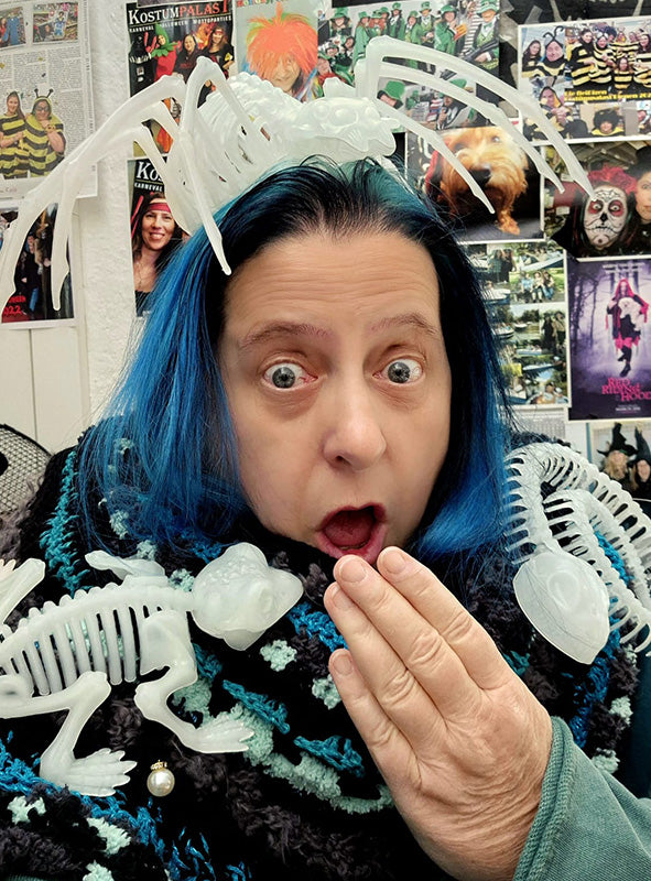 Eine Person mit blauen Haaren, die die nachtleuchtenden „Skeletttiere“-Halloween-Figuren trägt und von Spinnendekorationen umgeben ist, hält sich überrascht die Hand vor den Mund, während sie vor einer Wand steht, die mit Fotos und Postern bedeckt ist.