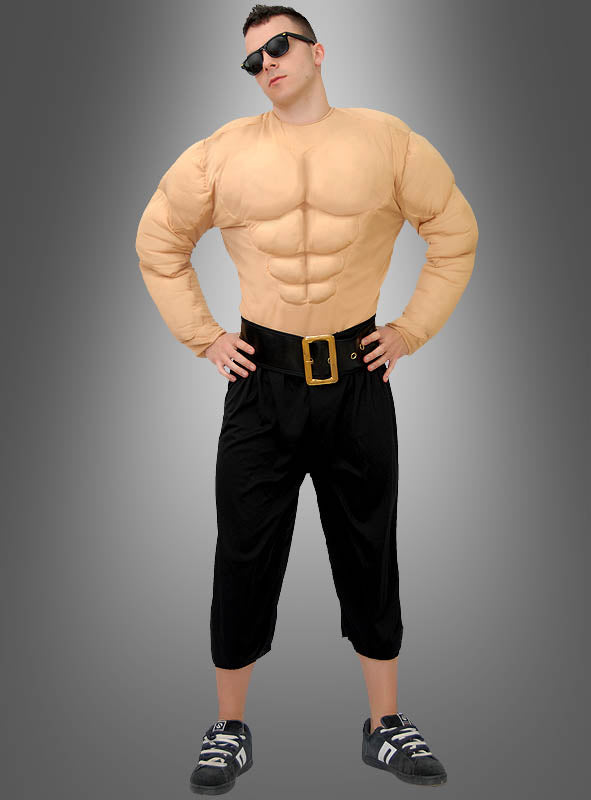 Eine Person, die eine schwarze Hose, schwarze Schuhe, das „Deluxe Muskelshirt Bodybuilder“-Kostüm – komplett mit einer großen Gürtelschnalle – und eine Sonnenbrille trägt, posiert mit den Händen in den Hüften vor einem grau verlaufenden Hintergrund.