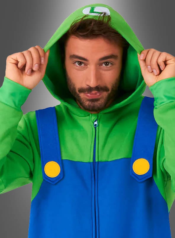 Eine Person, die den „Luigi-Overall für Erwachsene“ trägt – einen grün-blauen Kostüm-Hoodie mit gelben Knöpfen –, hält die Kapuze hoch, während sie vor einem grauen Hintergrund mit Farbverlauf steht. Damit eignet sich das Kostüm perfekt für den Fasching oder als Super-Mario-Kostüm.