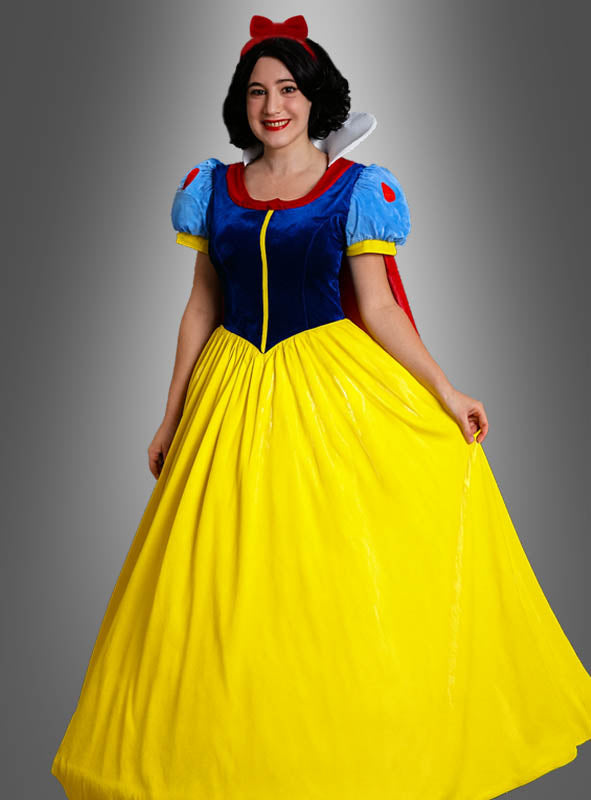 Eine Person, die das Märchenprinzessin-Schneewittchen-Kostüm trägt – bestehend aus einem blauen Oberteil, einem gelben Rock, einem roten Haarband mit Schleife und einem roten Umhang –, steht vor einem grauen Hintergrund.