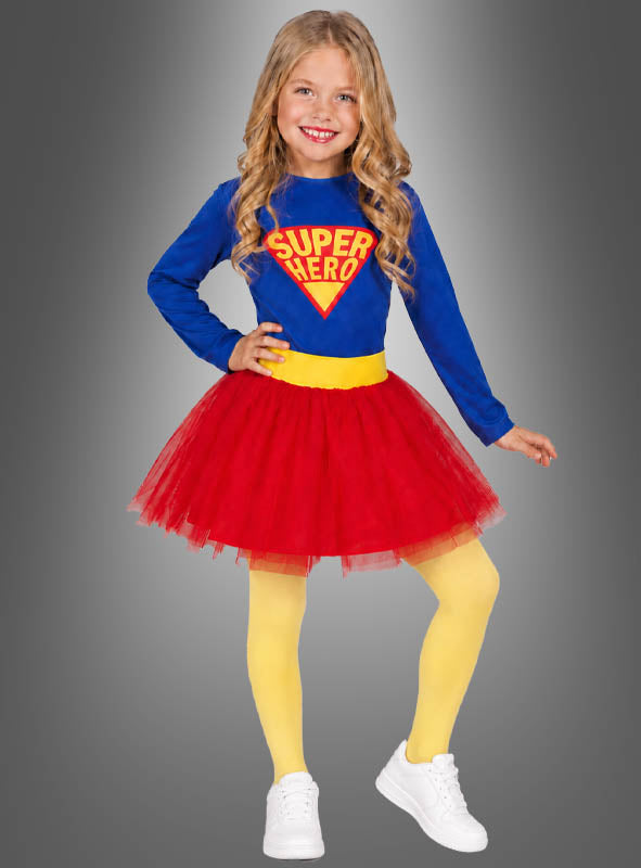 Ein kleines Mädchen steht lächelnd vor einem grauen Hintergrund und ist perfekt gekleidet in einem bezaubernden Superheldinnen-Kinderkostüm – bestehend aus einem blauen Superhelden-Shirt, einem roten Tüllrock, gelben Strumpfhosen und weißen Sneakers.