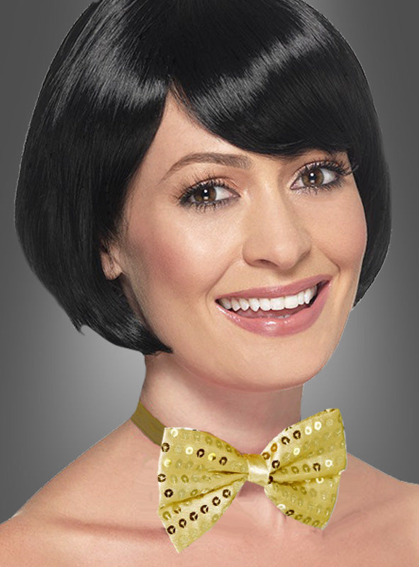 Eine lächelnde Person mit kurzen schwarzen Haaren trägt eine glänzende „Goldene Paillettenfliege“, die mit Pailletten verziert ist und sich vor einem grauen Hintergrund abhebt.