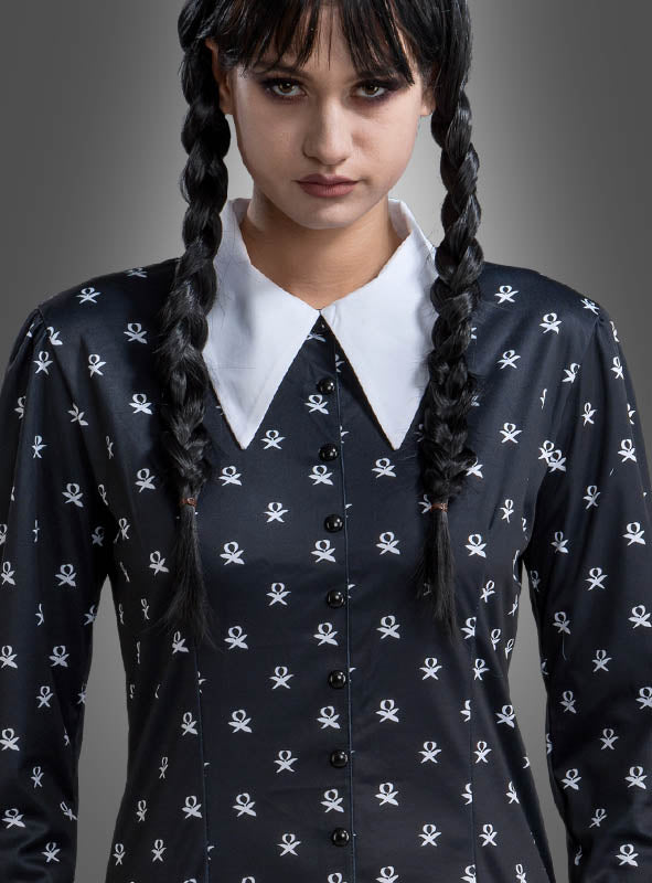 Eine Frau mit geflochtenen Haaren trägt das „Wednesday Damen Kleid schwarz-weiß“ – ein schwarzes Kleid mit weißen Totenkopfmustern und einem weißen spitzen Kragen –, das an das ikonische Wednesday-Kostüm aus der Addams Family angelehnt ist, und steht vor einem grauen Hintergrund.