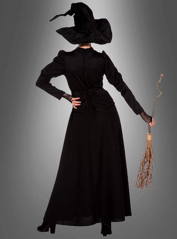 Eine Person trägt das „klassische schwarze Hexenkleid“ mit spitzem Hut, steht mit einem Besen in der Hand und dem Rücken zur Kamera vor einem grauen Hintergrund.