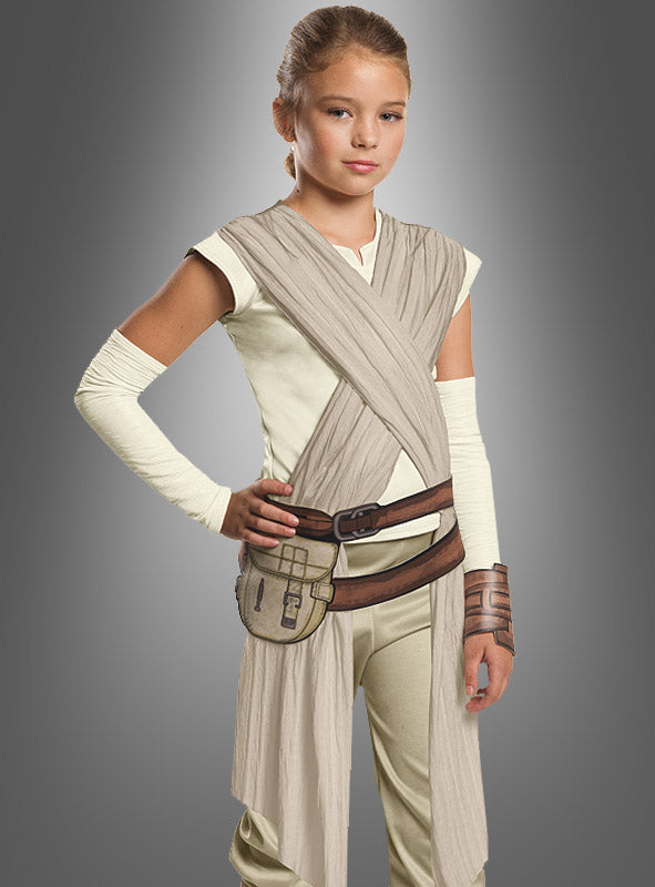 Ein junges Mädchen trägt das „Rey Star Wars Deluxe“-Kinderkostüm in Beige und Braun, das von der legendären Science-Fiction-Figur inspiriert ist, und steht selbstbewusst vor einem schlichten grauen Hintergrund.