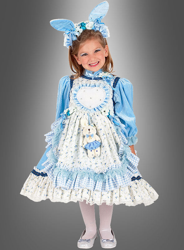 Ein kleines Mädchen steht lächelnd vor einem grauen Hintergrund und trägt das „Alice Deluxe“-Kinderkostüm – bestehend aus einem blau-weißen Rüschenkleid mit einer herzförmigen Schürze, einem Stirnband mit Hasenohren und weißen Strumpfhosen.