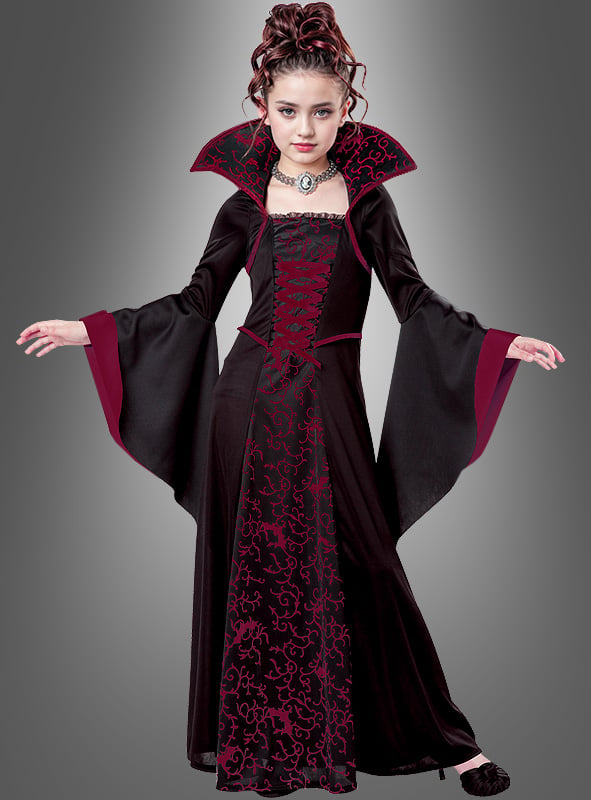 Ein junges Mädchen trägt das im Gothic-Stil gehaltene „Vampir-Königin“-Kinderkostüm „Mina“ mit hohem Kragen, ausgestellten Ärmeln und auffälligem rot-schwarz gemustertem Stoff und steht vor einem schlichten grauen Hintergrund. Dieses elegante Kostüm ist die ideale Wahl als Halloween-Kinderkostüm für alle, die den klassischen Vampir-Look lieben.