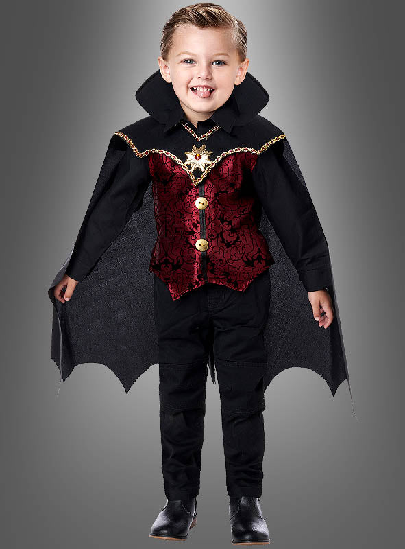 Ein kleines Kind trägt das „Edler Dracula“-Kostüm für Kinder mit auffälligen schwarzen und roten Details und einem wallenden Umhang und steht lächelnd vor einem schlichten grauen Hintergrund.